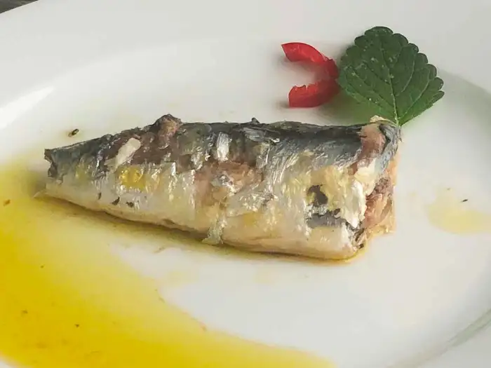 Sardinenstück in Öl mit Paprika und einem grünen Blatt auf einem weißen Teller