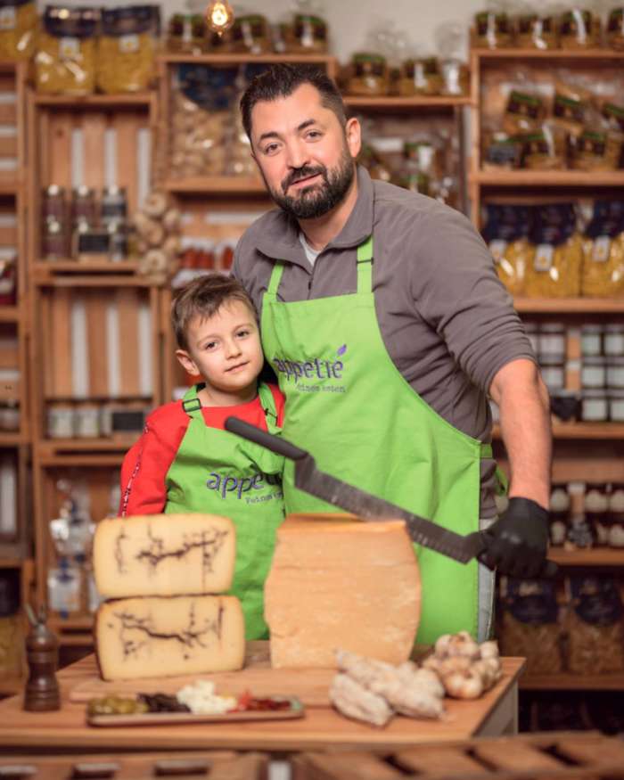 Inhaber Sergej Gricenko und sein Sohn in grünen Appetié-Schürzen hinter einer Auswahl an Käse und Feinkost